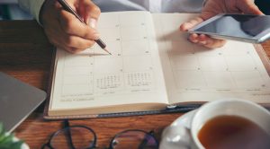 Kuvassa henkilö merkitsee kalenteriin kynällä. Kädessä on laskin ja pöydällä kahvikuppi ja silmälasit. Kuvituskuva