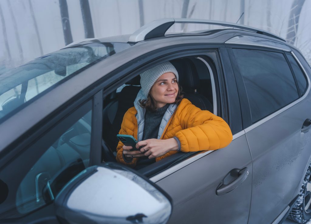 Nuori talvivaatteisiin pukeutunut nainen istuu autossa kännykkä käsissään. Hän nojaa kyynärpäällä avoinaisen ikkunan alareunaan ja katse on kääntynyt sivulle yläviistoon ulos autosta.