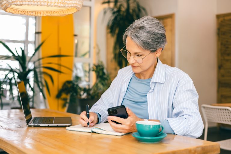Keski-ikäinen nainen työskentelee pöydän ääressä. Hänellä on edessään kannettava tietokone ja toisessa kädessään puhelin ja toisessa kynä. Pöydällä on kahvikuppi.