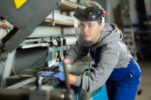 Nuori naistyöntekijä työskentelee suojakypärä päässä mekaanisen työstölaitteen ääressä.