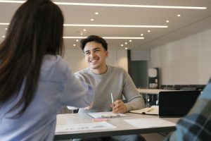 Nuori hymyilevä mieshenkilö kättelee pöydän yli naishenkilöä, joka on selin.