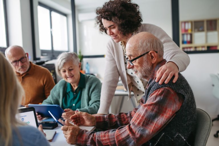 Kuvassa on ryhmä seniori-ikäisiä ihmisiä, jotka opettelevat tutorin avulla digilaitteiden käyttöä.