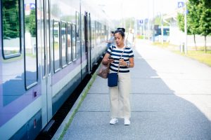 Kännykkä kädessä oleva aasialainen nainen on nousemassa junaan. Hänellä on kädessään kahvimuksi ja olalla kaksi laukkua.